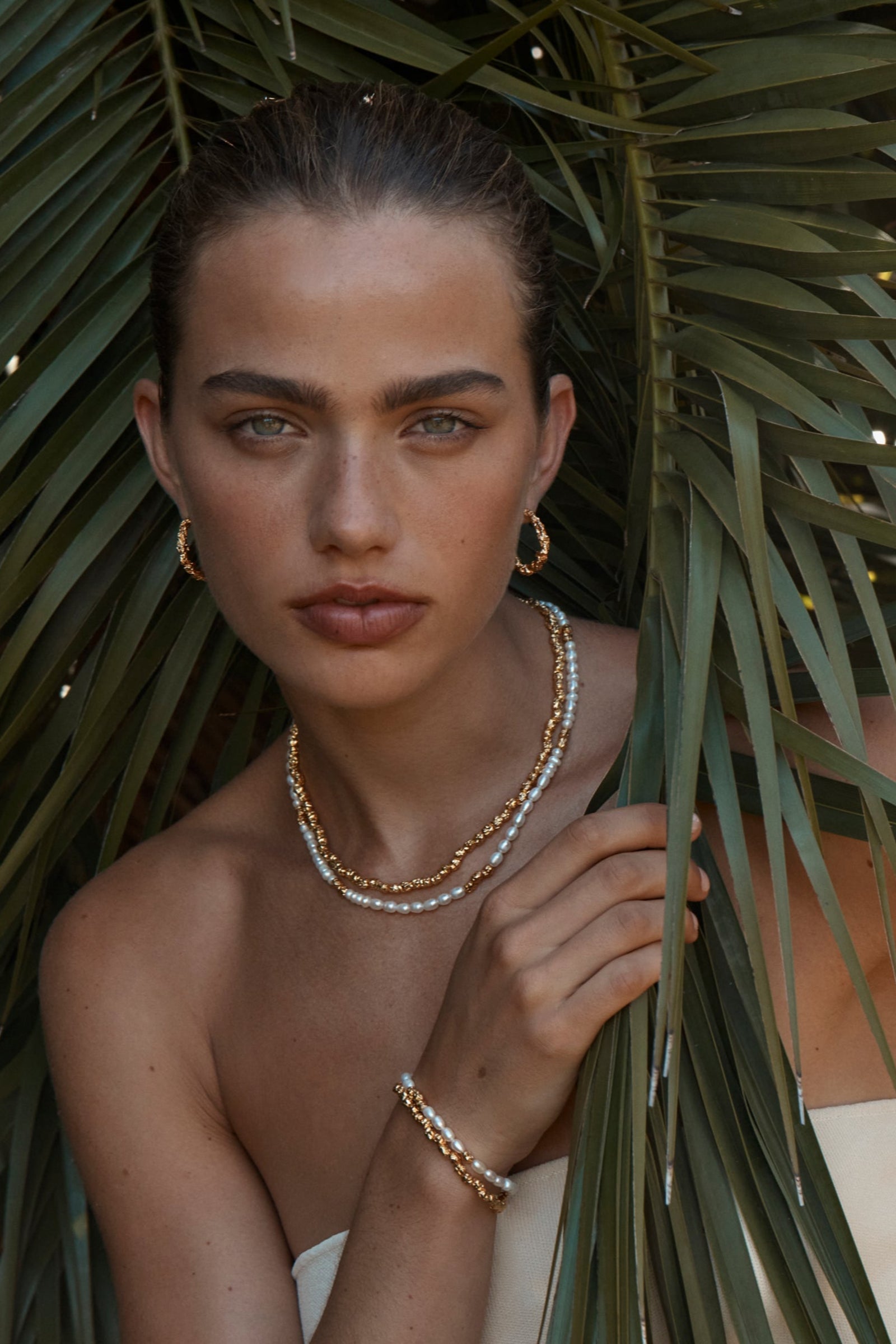 A person wearing a white dress and gold jewelry, including a double strand bracelet with pearls and gold textured pendants, posing among green foliage.