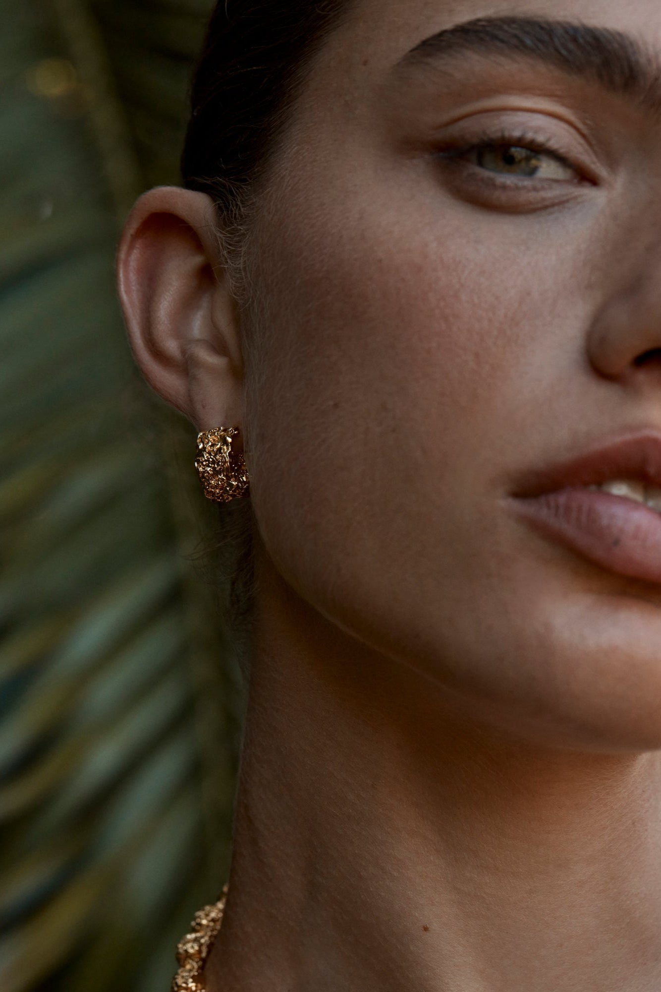 A person wearing a white tank top adorned with a gold-colored necklace and matching earrings, featuring a textured finish.