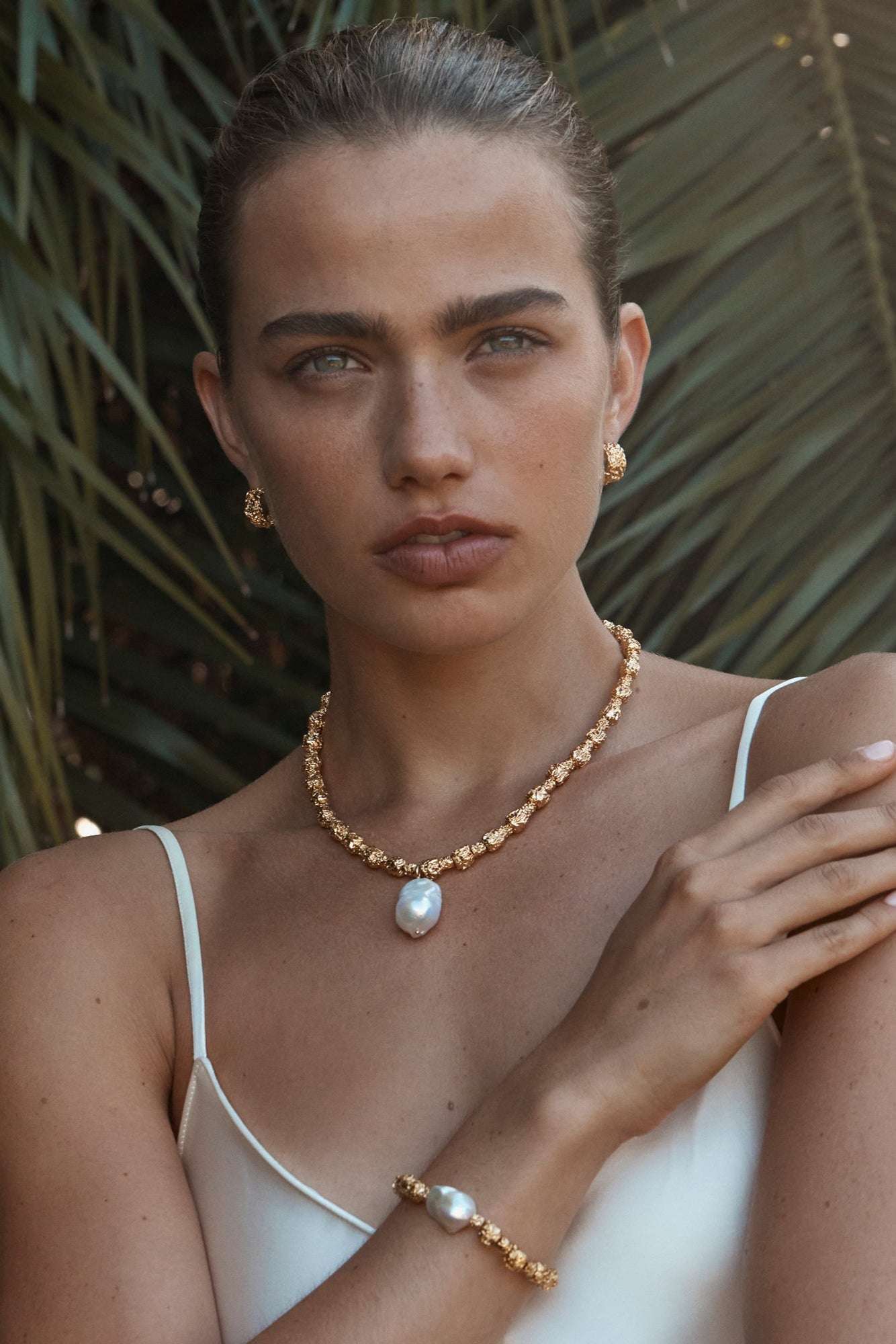 A person wearing a white tank top adorned with a large freshwater pearl necklace and matching earrings, set against a backdrop of green foliage.