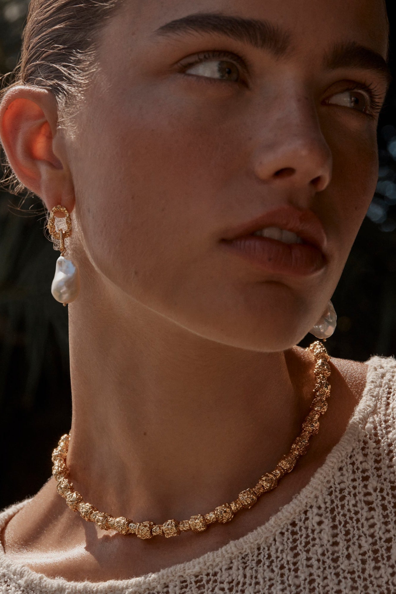 A person wearing a pair of gold-colored Peony earrings with freshwater pearls suspended beneath textured chain link pendants.