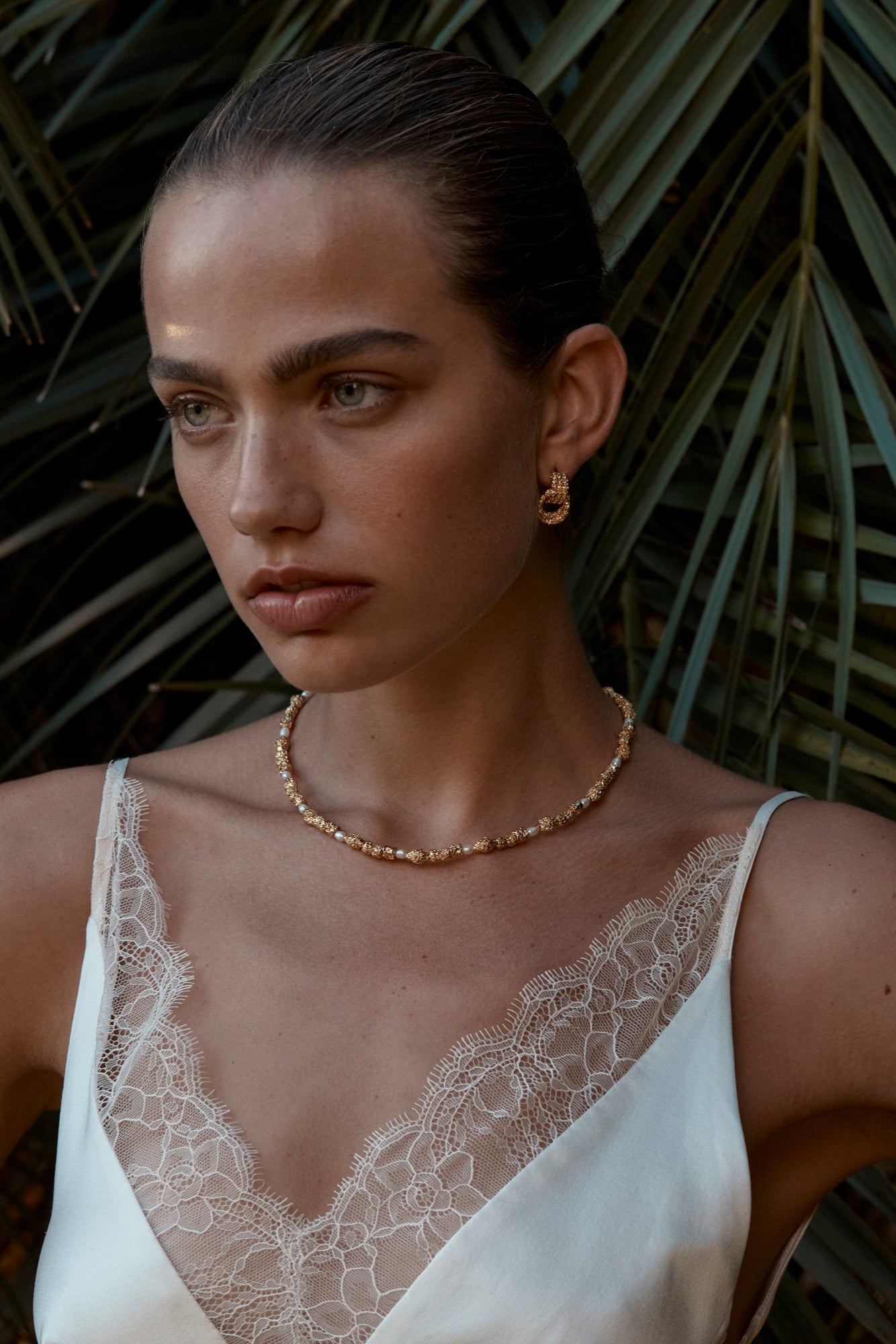 A person wearing a white top with lace details, showcasing gold-colored earrings and a matching bracelet.