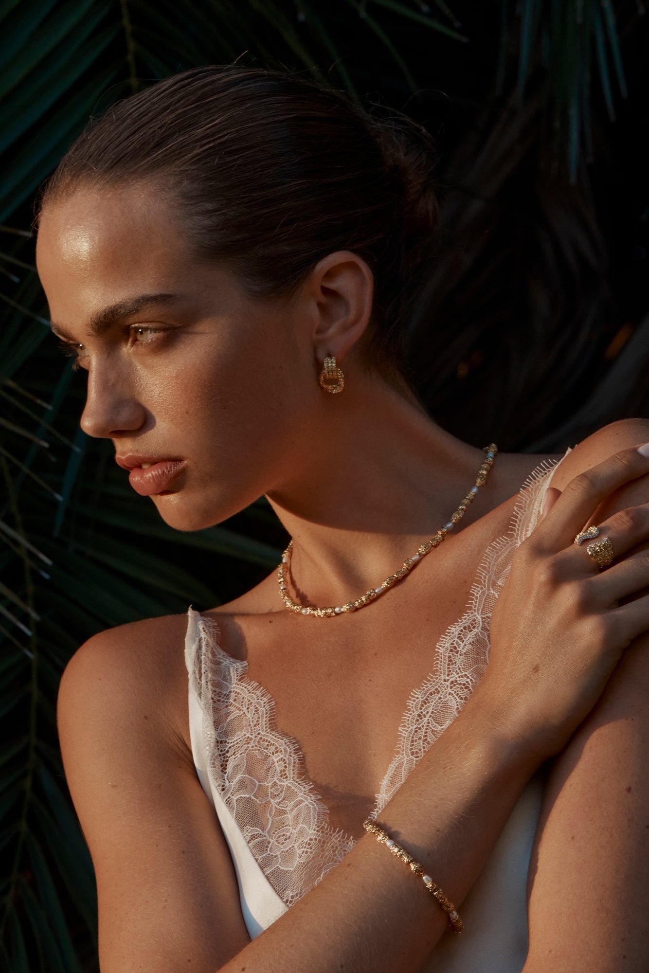 A person wearing a white top with lace details, showcasing gold-colored earrings and a matching bracelet.