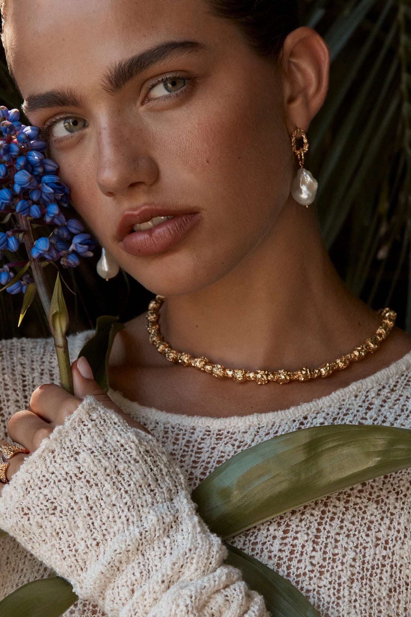 A person wearing a pair of gold-colored Peony earrings with freshwater pearls suspended beneath textured chain link pendants.