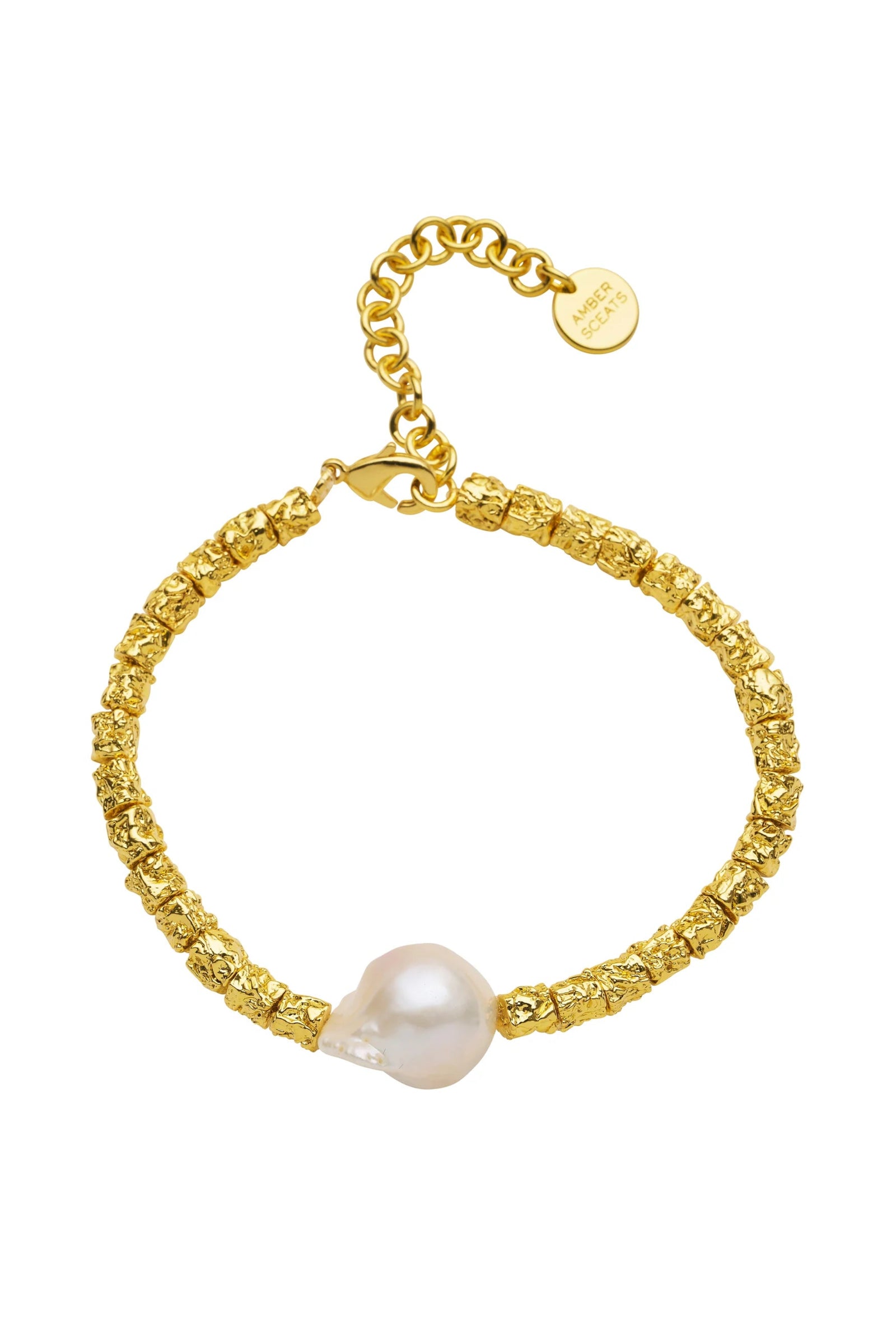 A person wearing a beige dress and gold jewelry, including a bracelet and necklace, with a textured background.