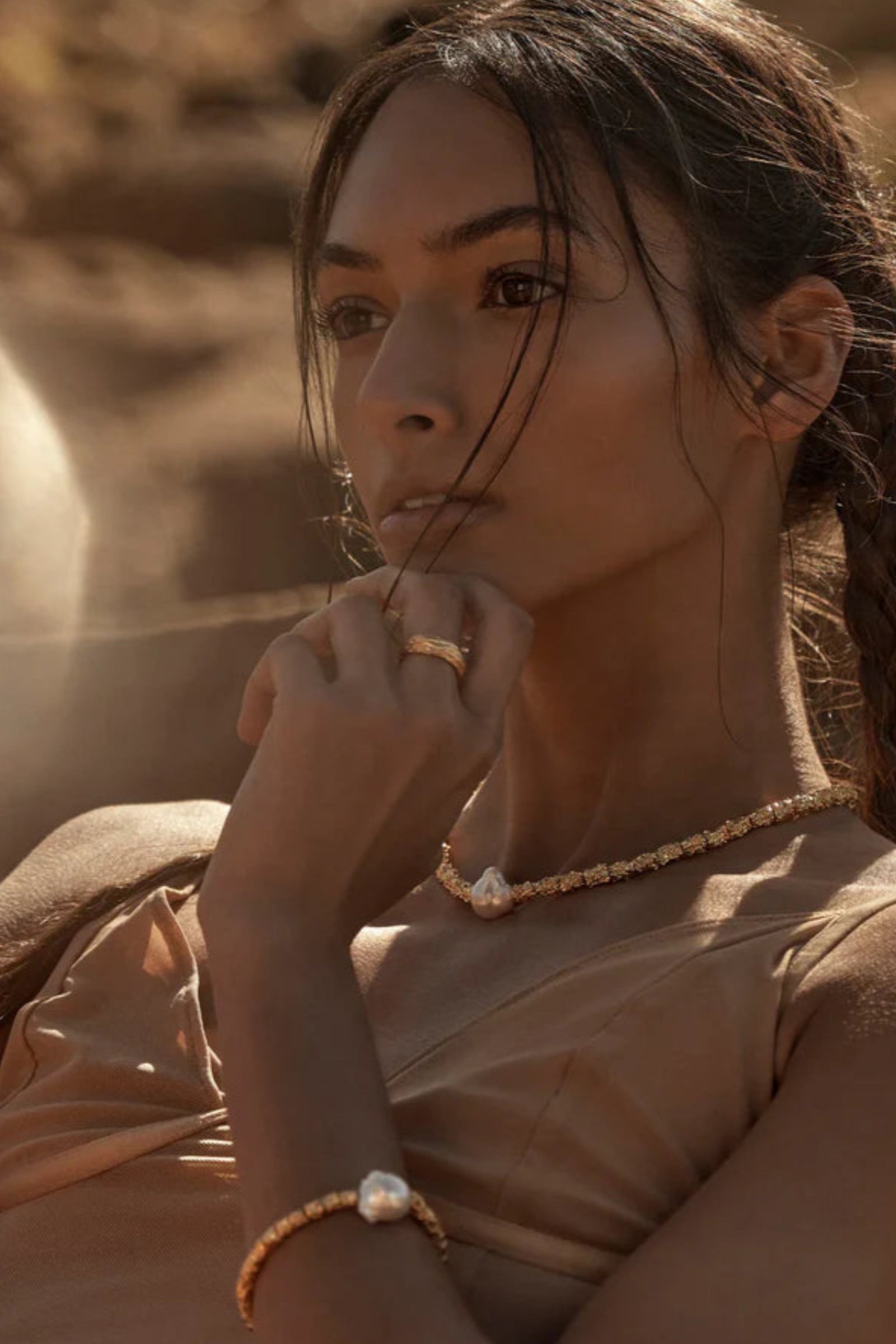 A person wearing a beige dress and gold jewelry, including a bracelet and necklace, with a textured background.