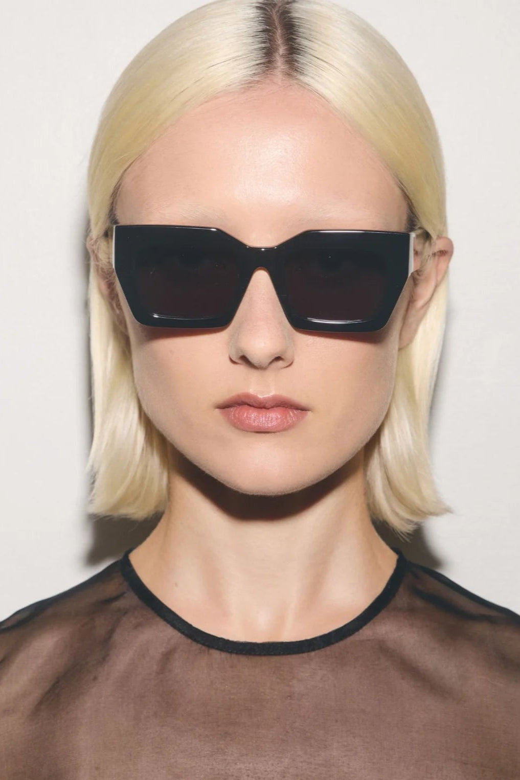 Person wearing dark sunglasses and a sheer brown top against a plain background