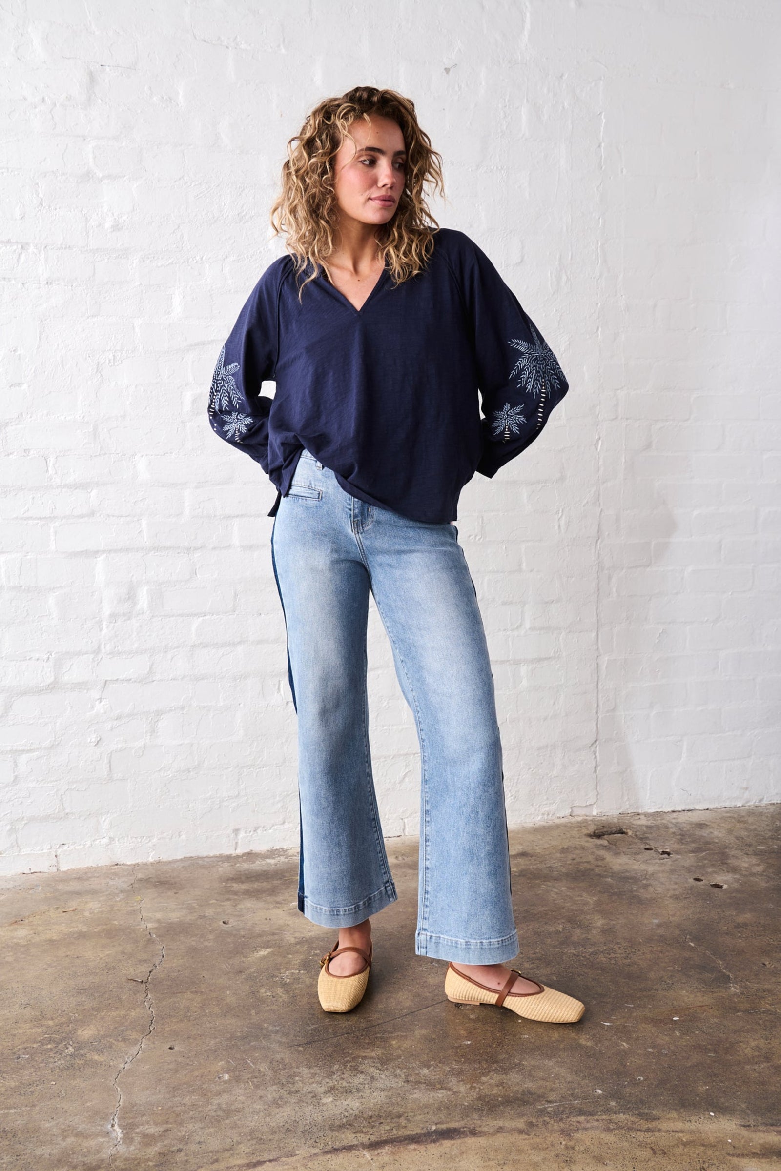 Woman wearing a navy blue blouse and light blue jeans standing against a white brick wall.