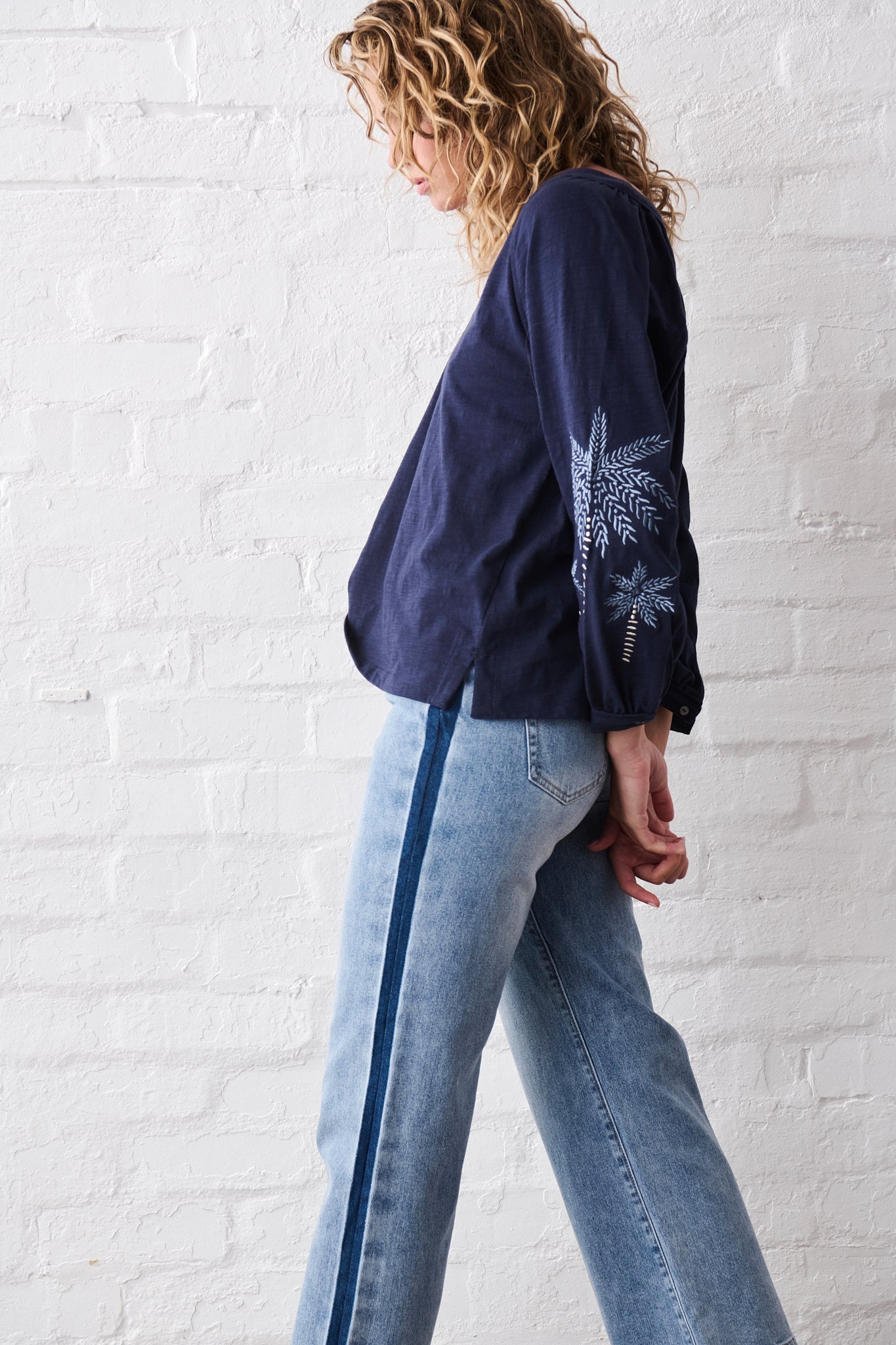 Woman wearing a navy blue blouse and light blue jeans standing against a white brick wall.