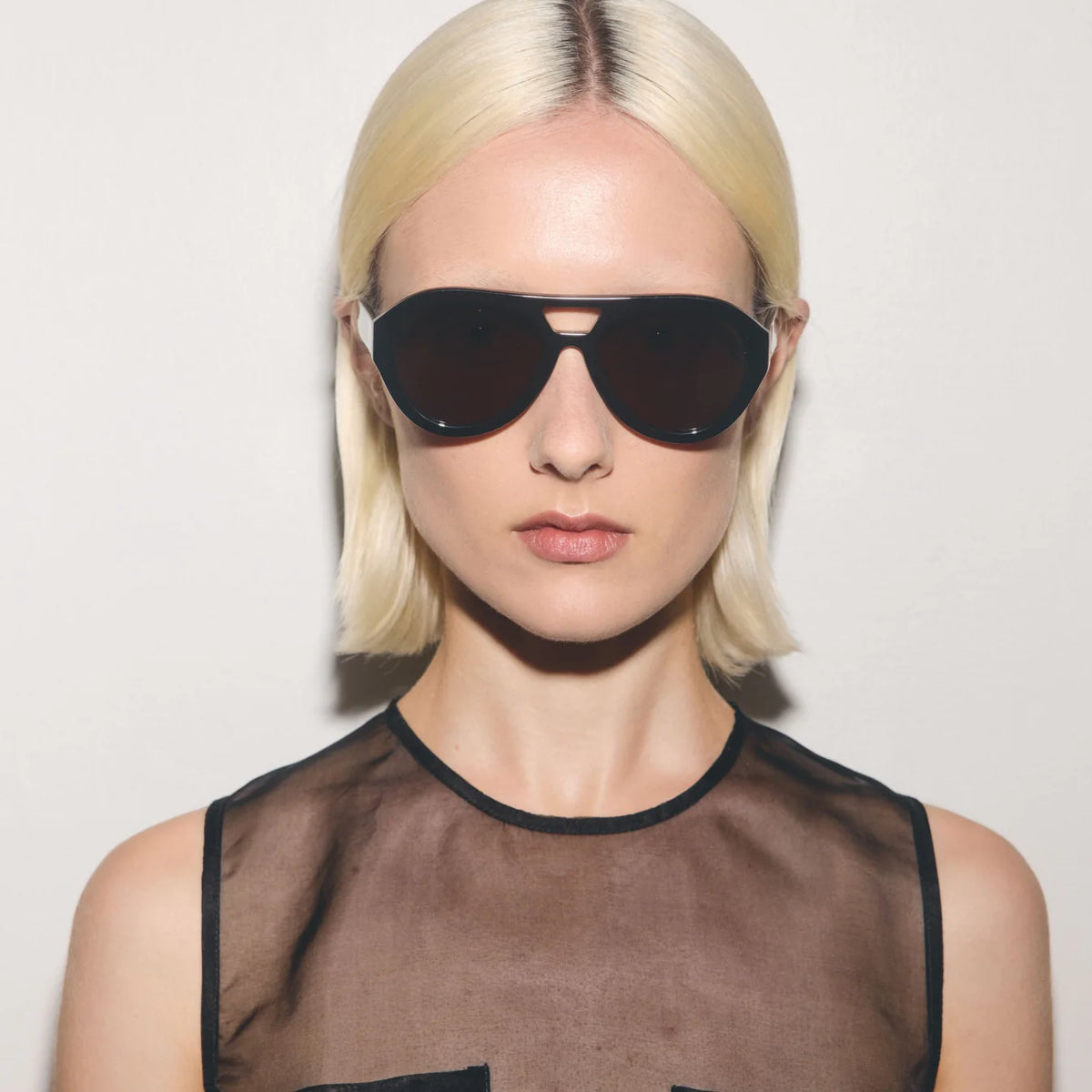 Person wearing black sunglasses and a sheer top against a plain background