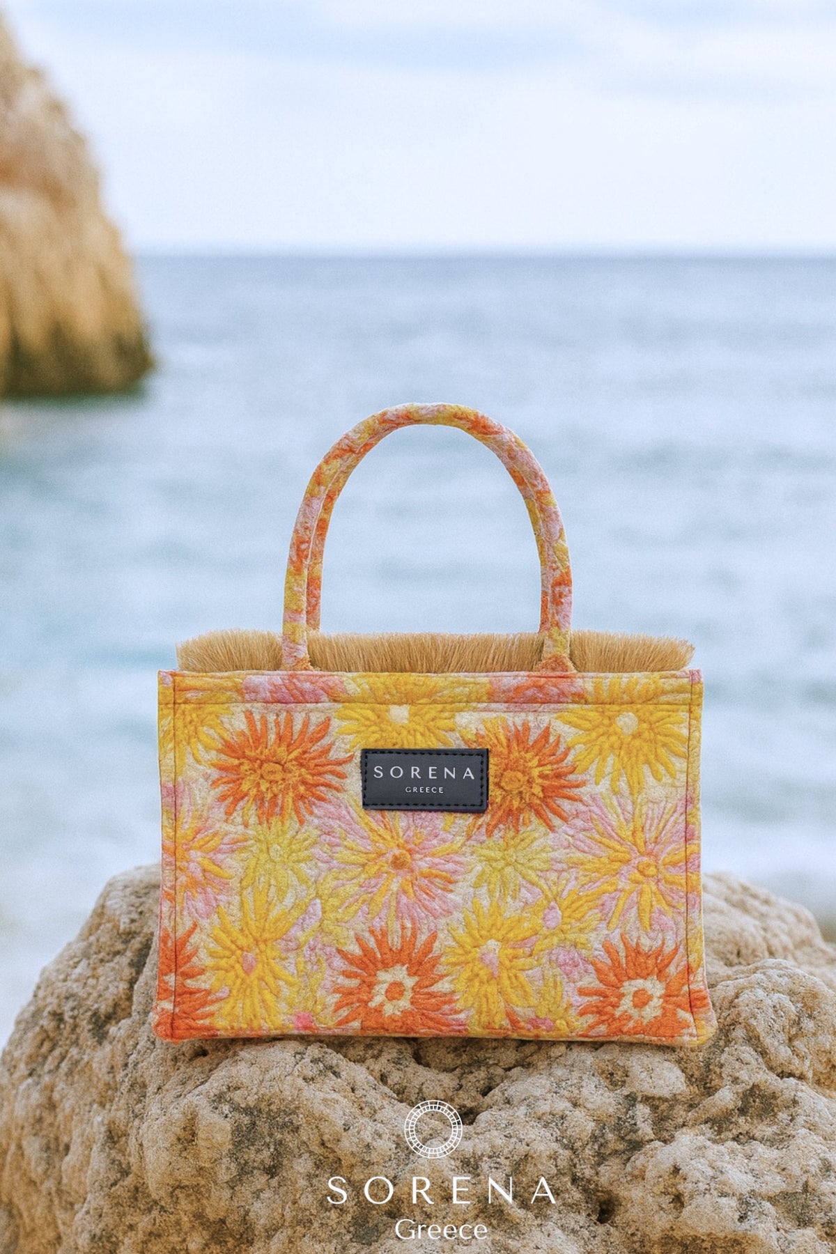 Floral patterned bag on a rock with ocean in the background, featuring the brand 'Sorena'.
